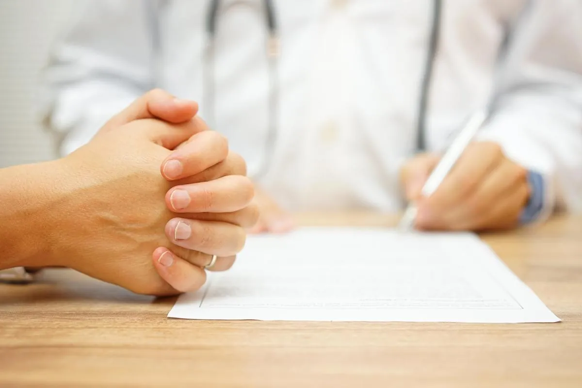 Patient with clasped hands across a desk from a doctor writing on a form